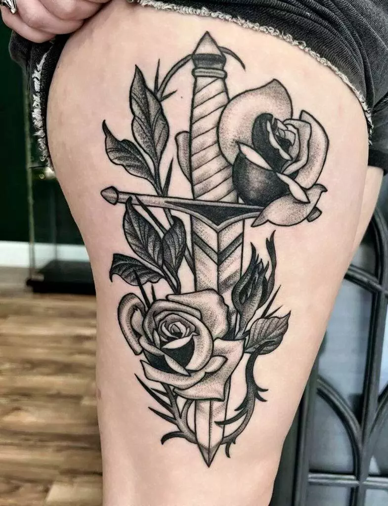 Black and grey tattoo of a dagger piercing through two roses with leaves, located on an upper thigh.