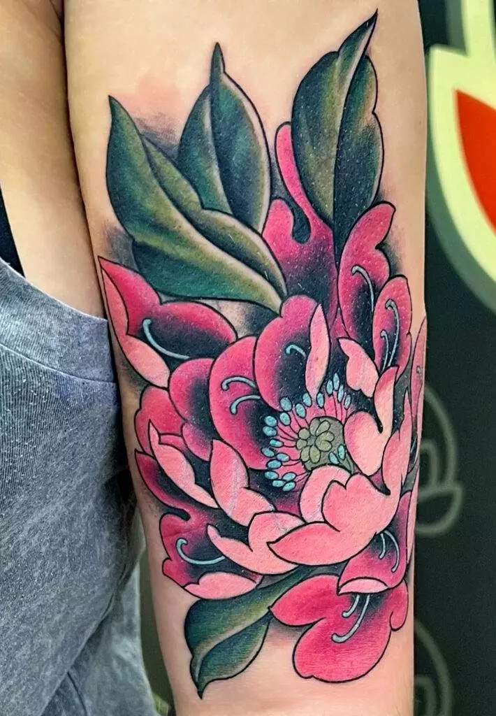 A large tattoo featuring a vivid pink peony with green leaves is displayed on a person's upper arm.