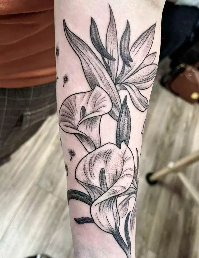 A forearm tattoo featuring detailed black and gray flowers and leaves against a background of scattered seeds.