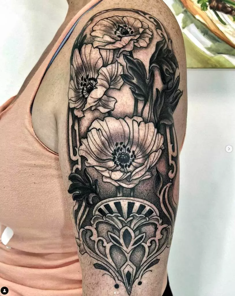 A person's upper arm with a detailed black and gray tattoo featuring large blooming flowers and intricate ornamental designs.